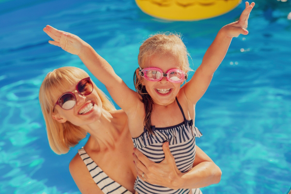 Woman and child in swimsuits smile and pose in a pool managed by a renowned pool service company, with the child wearing goggles and raising her arms.