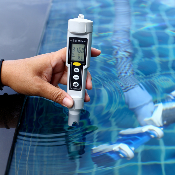 A hand holds a digital salt meter above a swimming pool, displaying readings on its screen.
