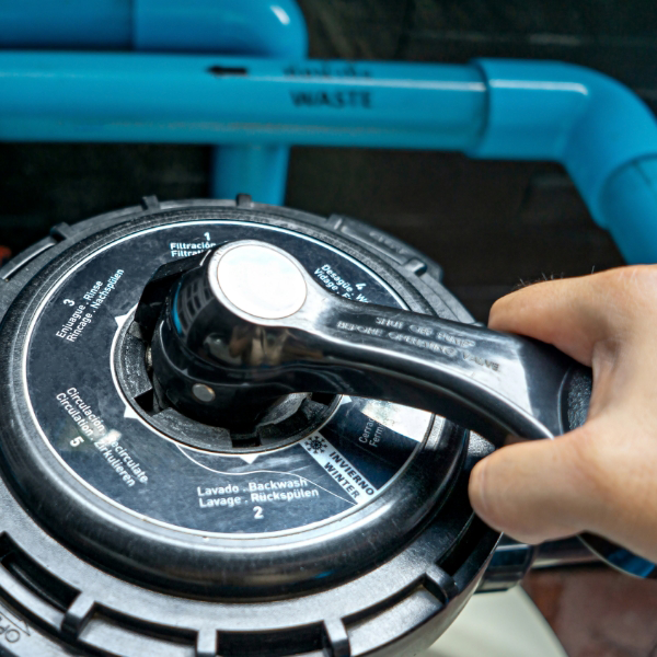 Hand adjusting a pool filter valve with directional control and multilingual labels; blue plumbing pipes visible in the background.